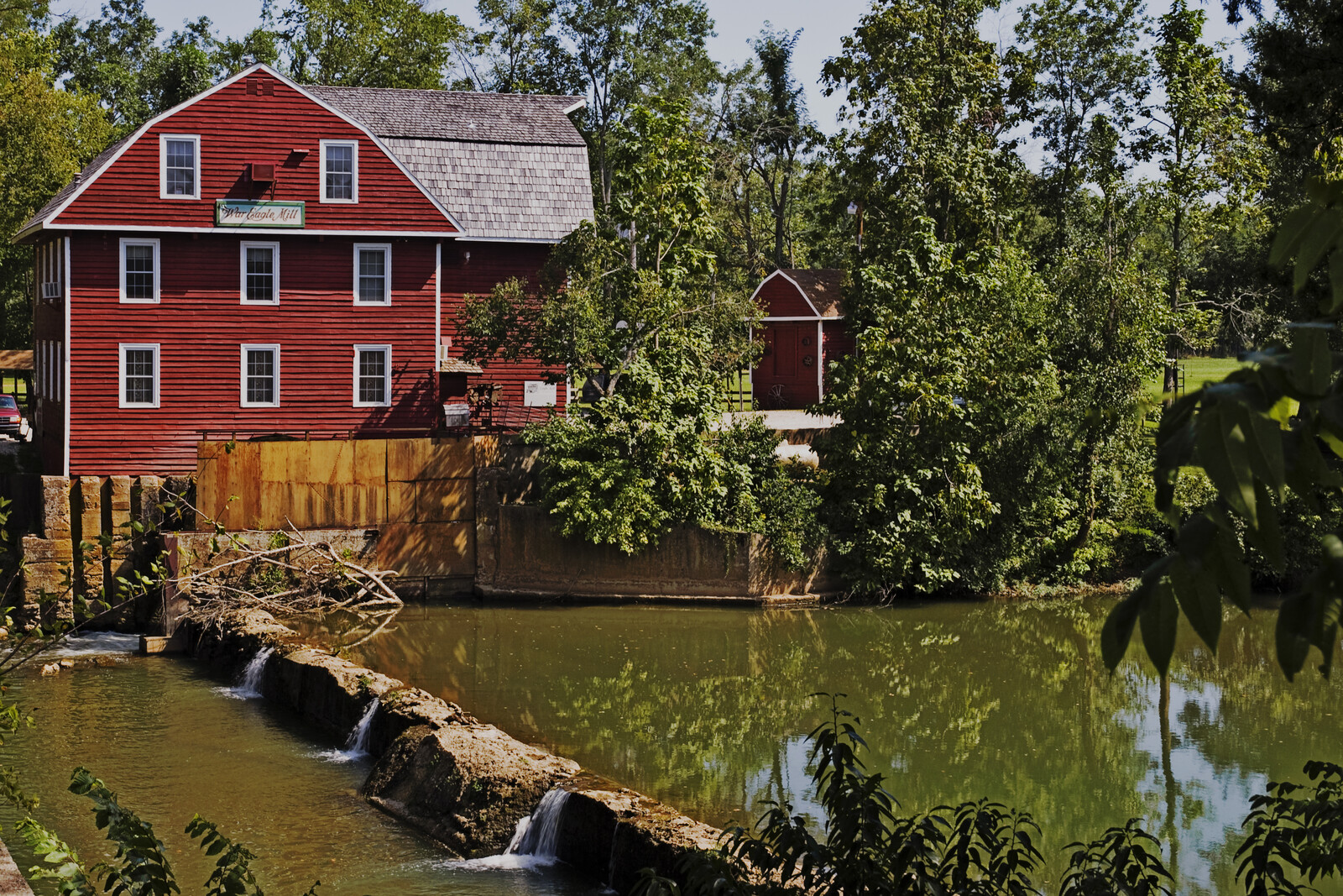 The red, white-trimmed War Eagle Mill rises beside a low stone dam and green water on War Eagle Creek, surrounded by trees.